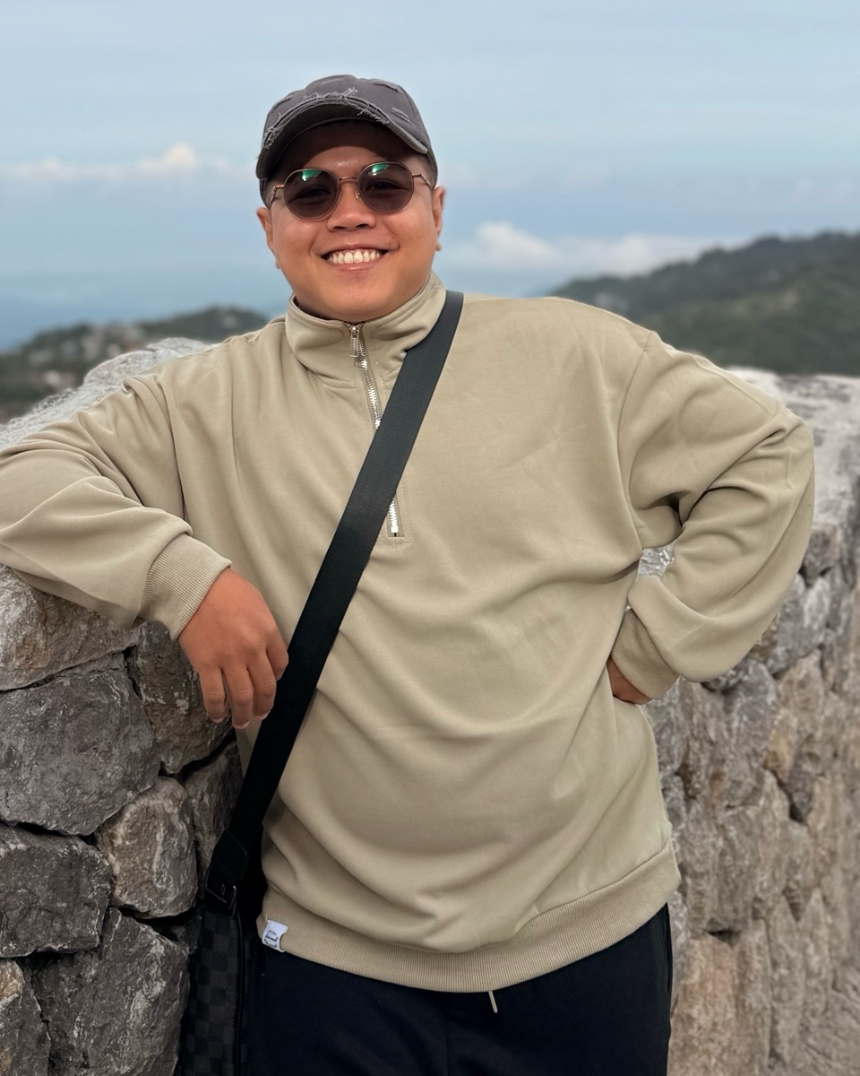 Earnest John Gungon, smiling, in a cap and sunglasses, leaning on a stone wall with hills behind him.